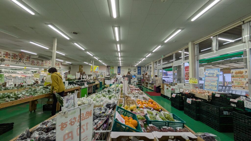 地元の産直センターの内部、緑色の床と照明が照らされ、さまざまな新鮮な野菜や果物が並んでいる。買い物客が商品を選んでいる様子が見られる。