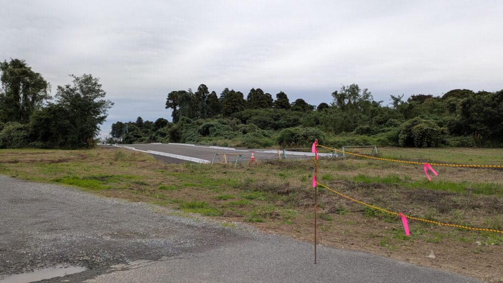 新しい道路工事中の風景。左側に整地された土地と、右側に道が見える。背景には緑豊かな樹木が広がっている。