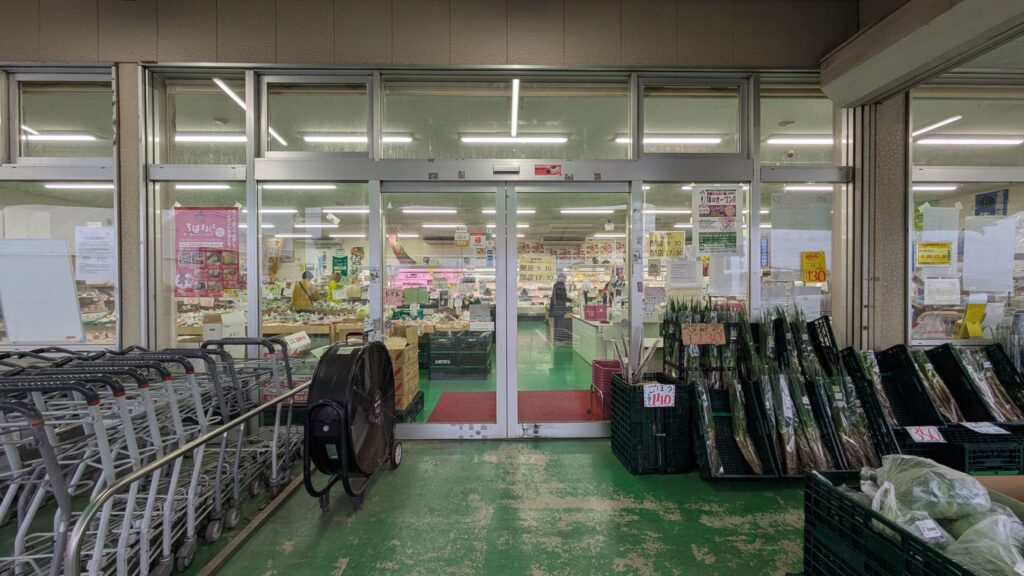 入口のガラスドアと緑色の床が見える産直センターの外観。左側にはカートと大きな扇風機が置かれ、右側には野菜が入った黒いバスケットが並んでいる。