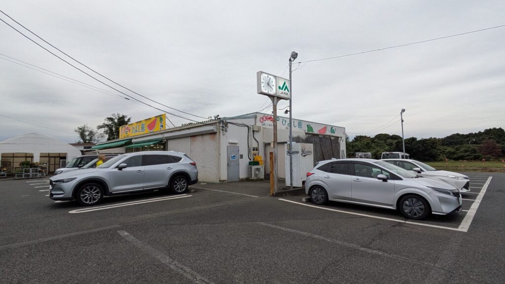 JA富里市産直センターひよし館の外観、駐車場に数台の車が停まっている。時計が見える。