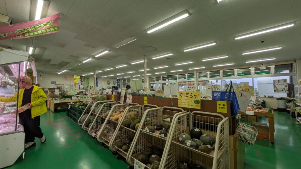 産直センターの内部風景、鮮やかな緑の床が特徴的。左側には冷蔵ケースと野菜が並び、右には買い物客が見える。