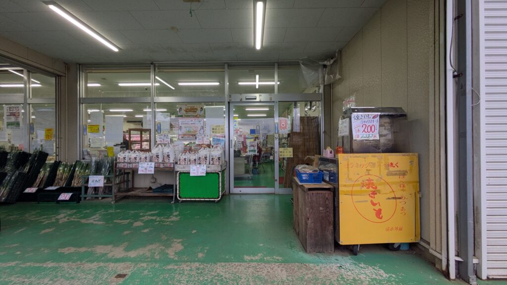産直センターの入り口の外観。店内は明るく、商品のポスターが掲示されている。緑の床が特徴的で、入り口の右側には焼き芋販売の黄色いスタンドが設置されている。