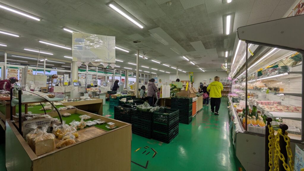 写真はJA富里市産直センター2号店の店内の様子を示しており、緑色の床と多様な商品が並ぶ陳列棚が見える。買い物客が商品を選んでいる風景が広がっている。