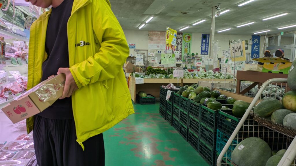 店内で食材を選ぶ人物が黄色のジャケットを着ている様子。周囲には新鮮な野菜が並ぶ農産物直売所の内部。