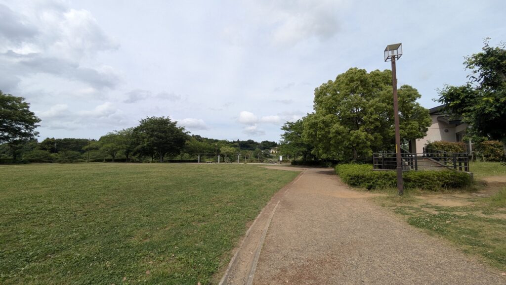 広々とした公園の風景、緑の芝生と山々が見える。散歩道があり、周囲には木々や施設が点在している。