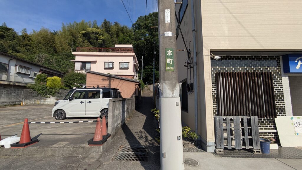 住宅地の道路の風景で、白い車と駐車用コーンが見える。奥にはピンクの建物があり、その周りには緑がある。