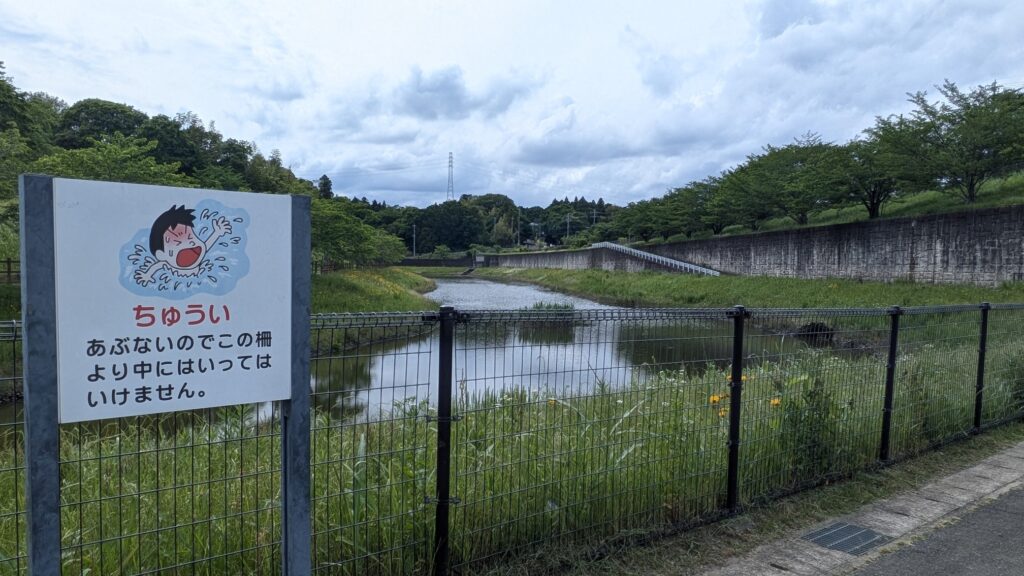 川の近くに設置された禁止標識があり、水に入ってはいけないことを警告している。背景には緑豊かな景色と雲が広がっている。
