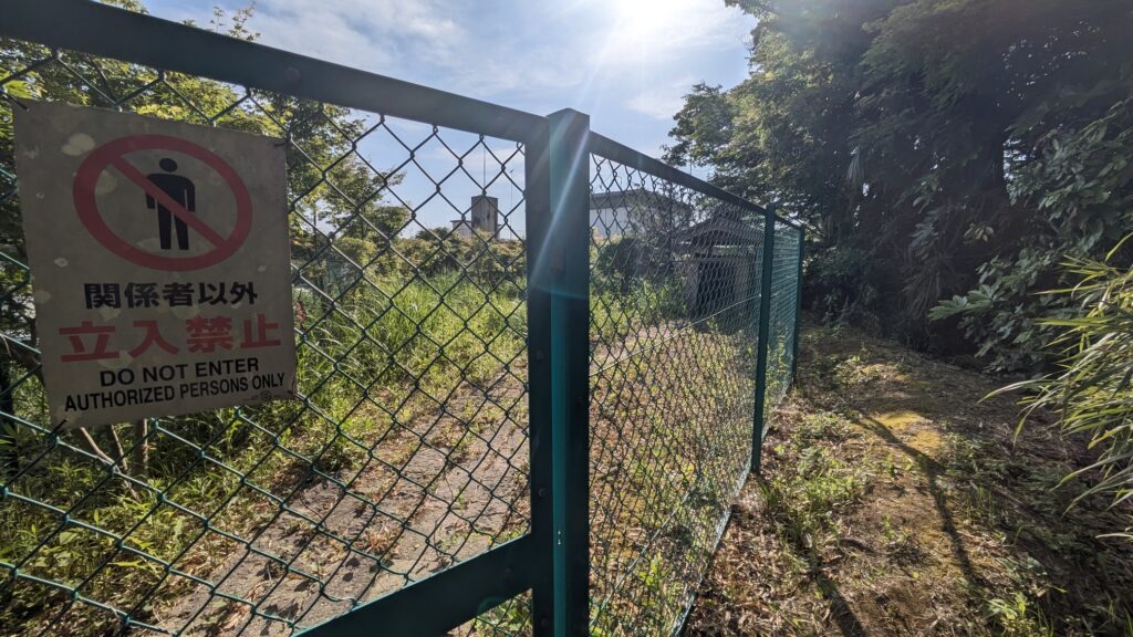 フェンスの前に立てられた立入禁止のサインがある風景。背後には青空と緑の植物が見え、太陽の光が当たっている。