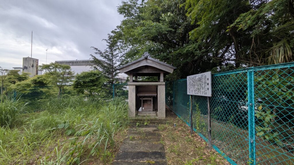 緑に囲まれた小さな祠がある風景。手前には舗装された道があり、背後には高い草や樹木が見える。周囲にはフェンスが設置され、奥には建物の一部が見える。