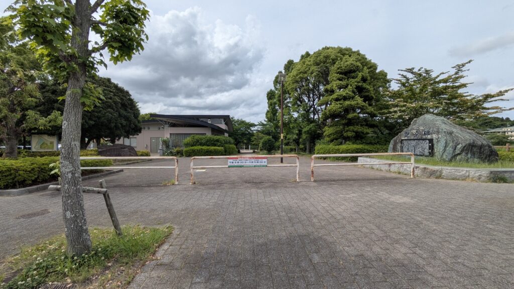 公園の入口にあるバリアと案内板が見える風景。周囲には緑の木々があり、雲の多い空が広がっている。