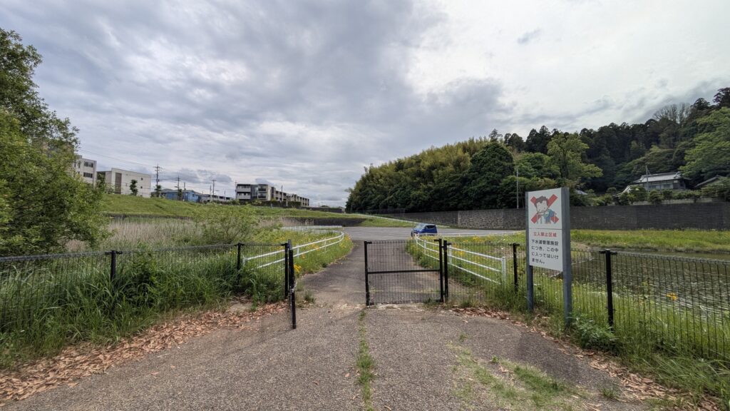公園の入り口の風景で、緑の草が生い茂り、フェンスで囲まれた道が見える。液体のある場所と木々が背景にあり、標識が立っている。