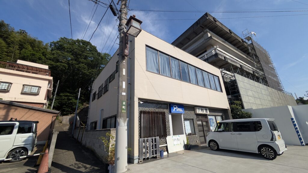 建物の外観、道路沿いにあり、前に白い車が停まっている。左側には他の建物と緑の木々が見える。