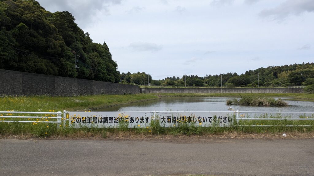 池と緑の草地が広がる風景。白いフェンスに「この場所は調整池があります」や「大雨時は立ち入らないでください」と書かれた看板が見える。