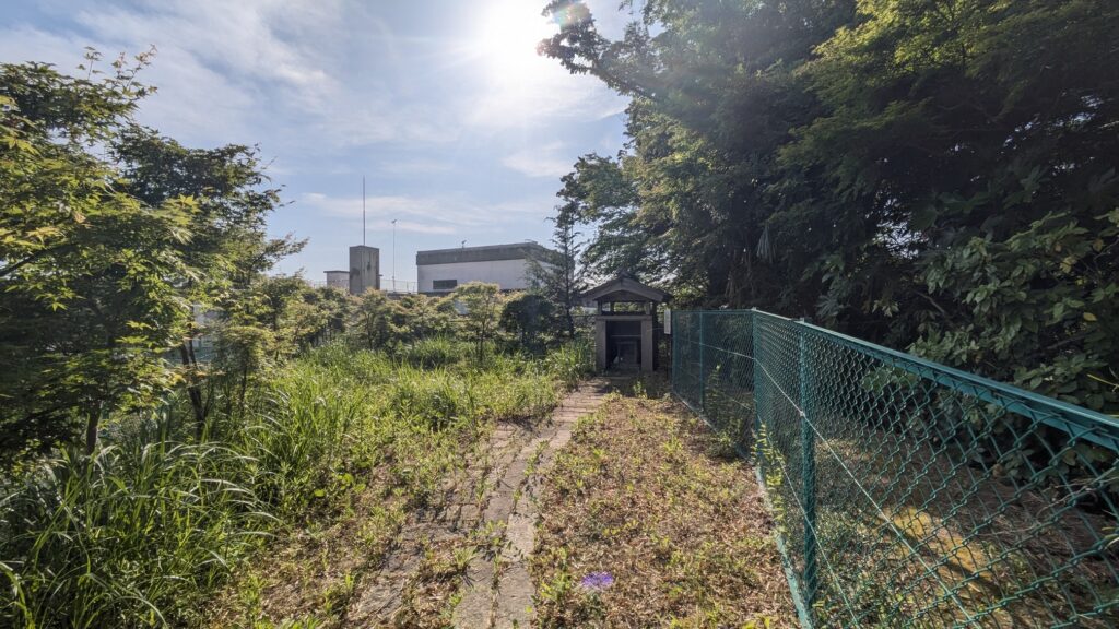 緑に囲まれた細い小道とフェンスがある風景。遠くに建物が見え、晴れた空の下で自然が広がっている。