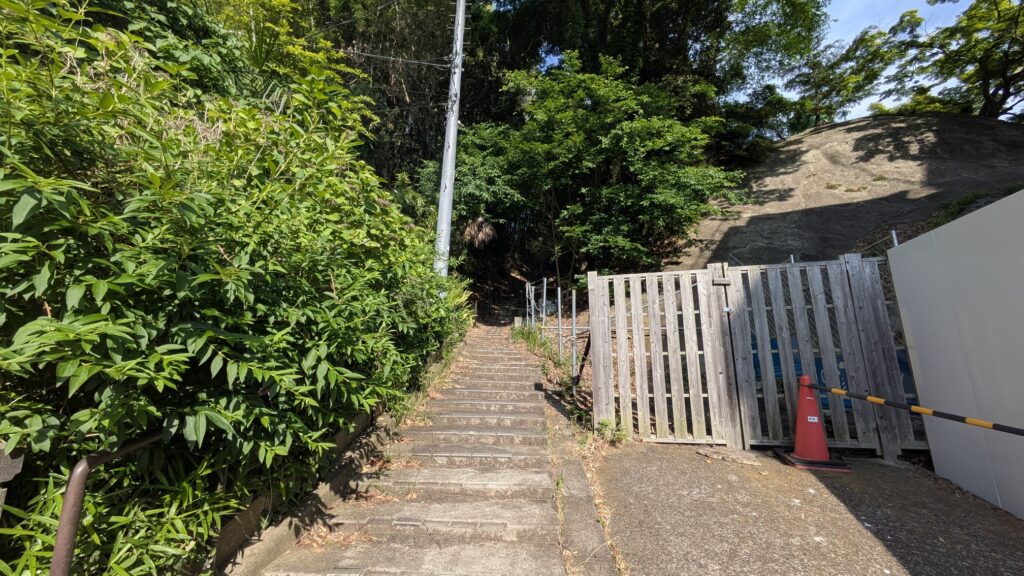 階段を上る緑に囲まれた道の風景。左側には木の柵と赤いコーンが見える。