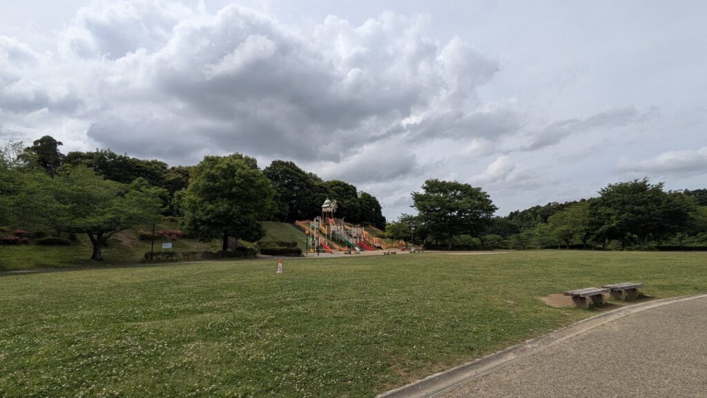 緑豊かな公園の広場に子供用の滑り台がある景色。空には雲が広がっている。