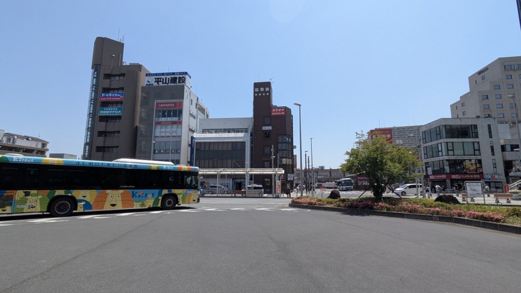 バスと商業ビルが並ぶ都市の風景。青空の下で、舗装された道路と植え込みが見える。