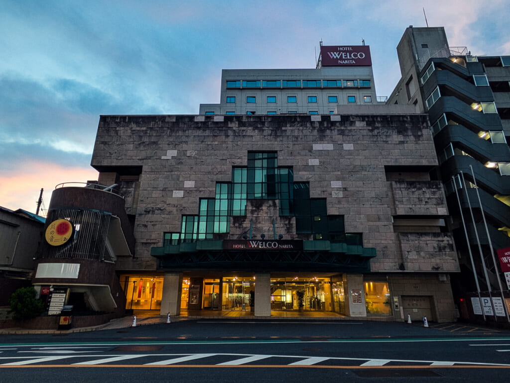 2026年10月撮影・成田市花崎町のホテルウェルコ成田 夕暮れの正面外観と階段状のガラス面|営業中